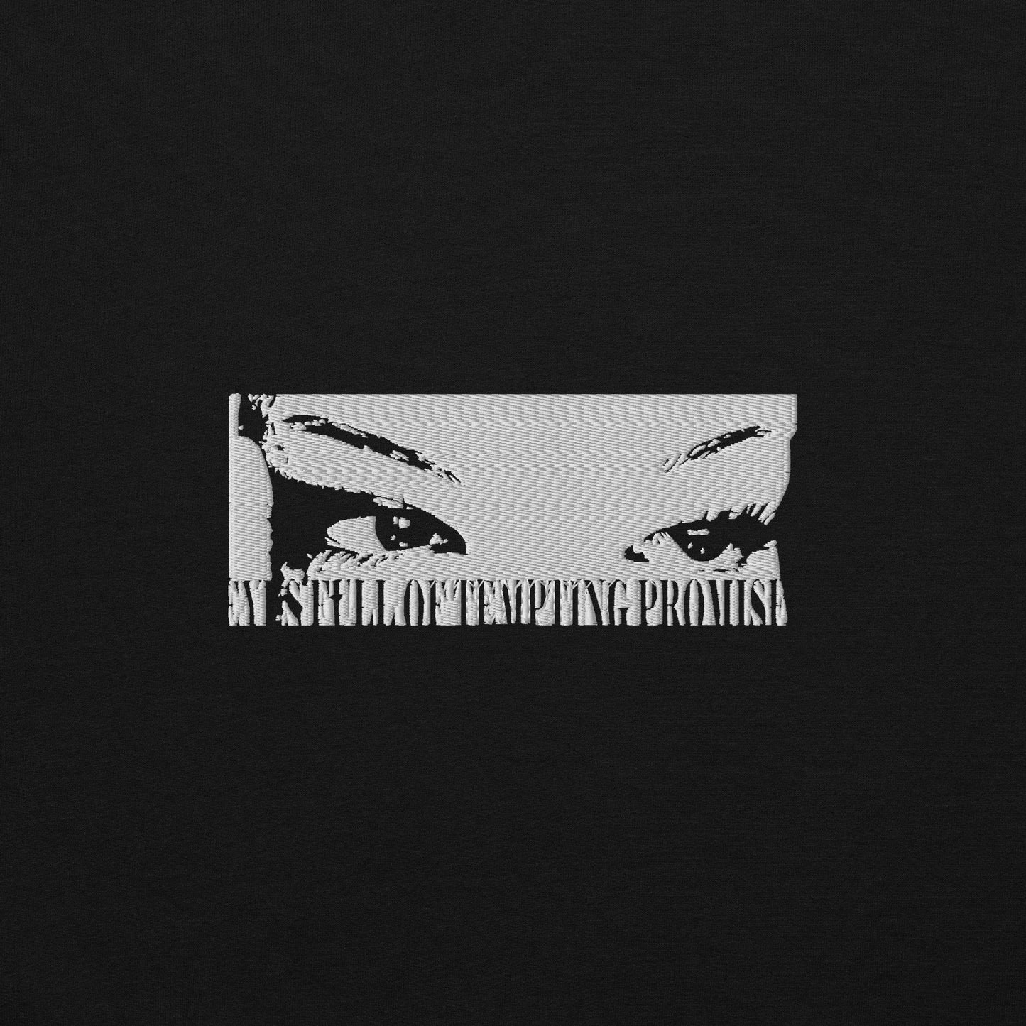 A white graphic stitched on a piece of fabric. The design is of a females eyes and there is a black stitched text underneath the graphic saying “Eyes full of tempting promises”