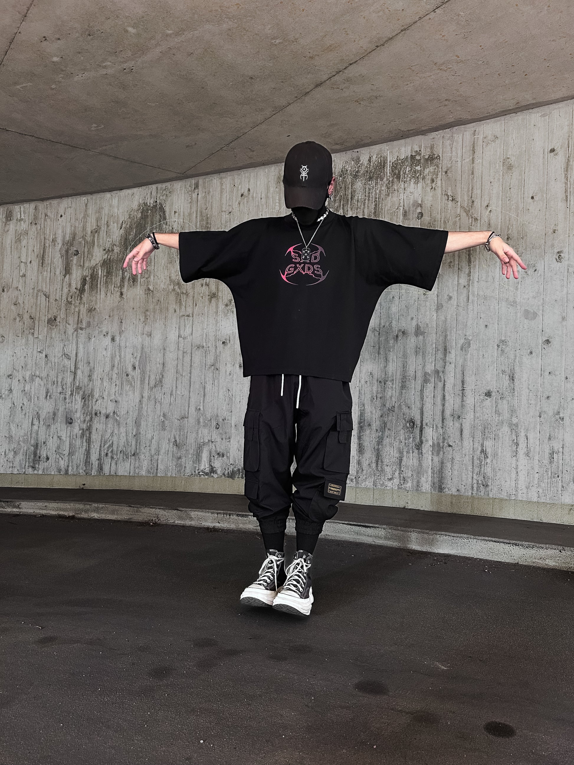 Person standing under a concrete structure wearing oversized black clothing with graphics and text on the shirt, black pants with pockets, and black and white sneakers. They are also wearing a black cap and have their arms extended out to the sides.