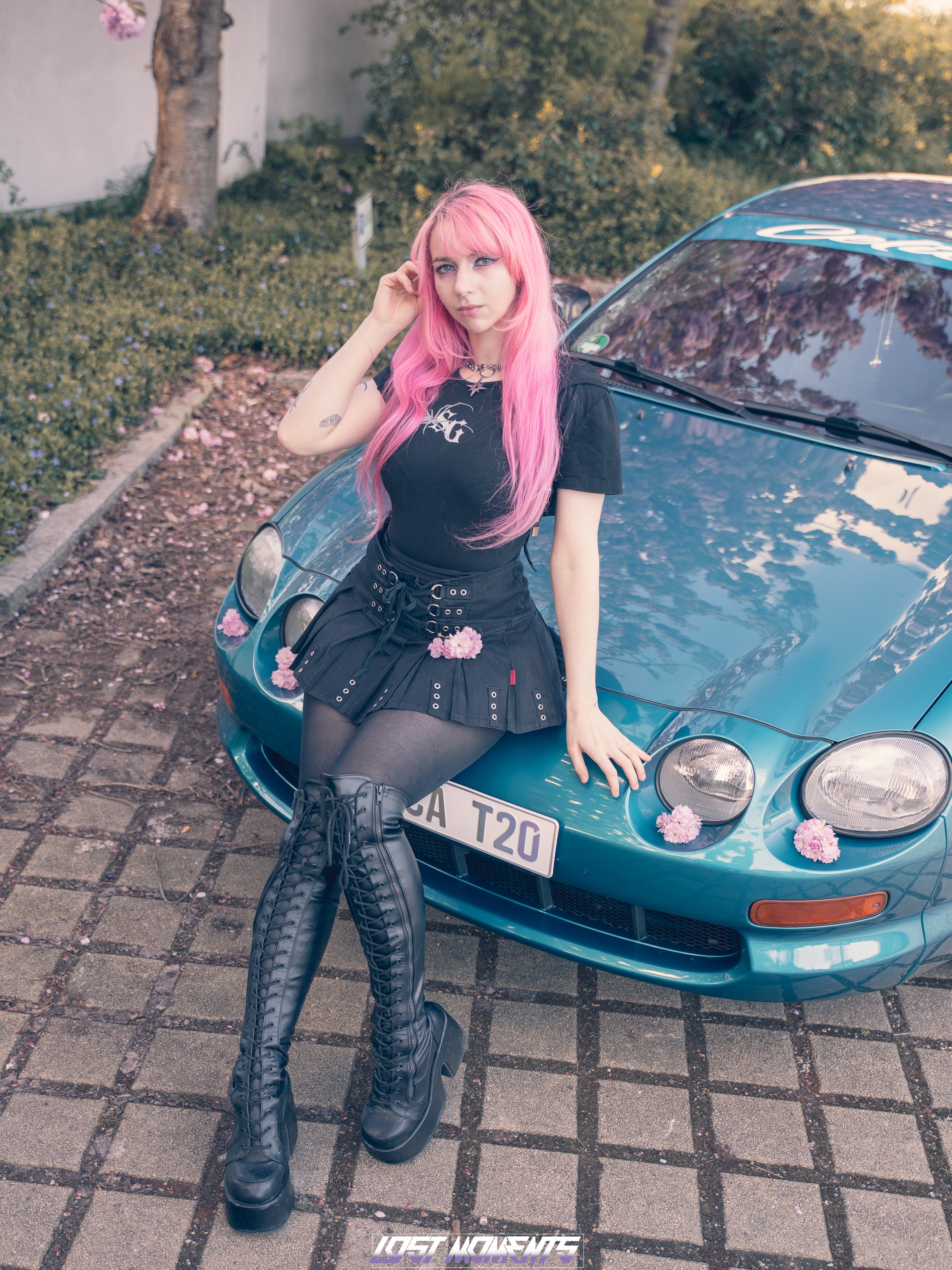 Person with pink hair leaning against a blue car on a paved area.