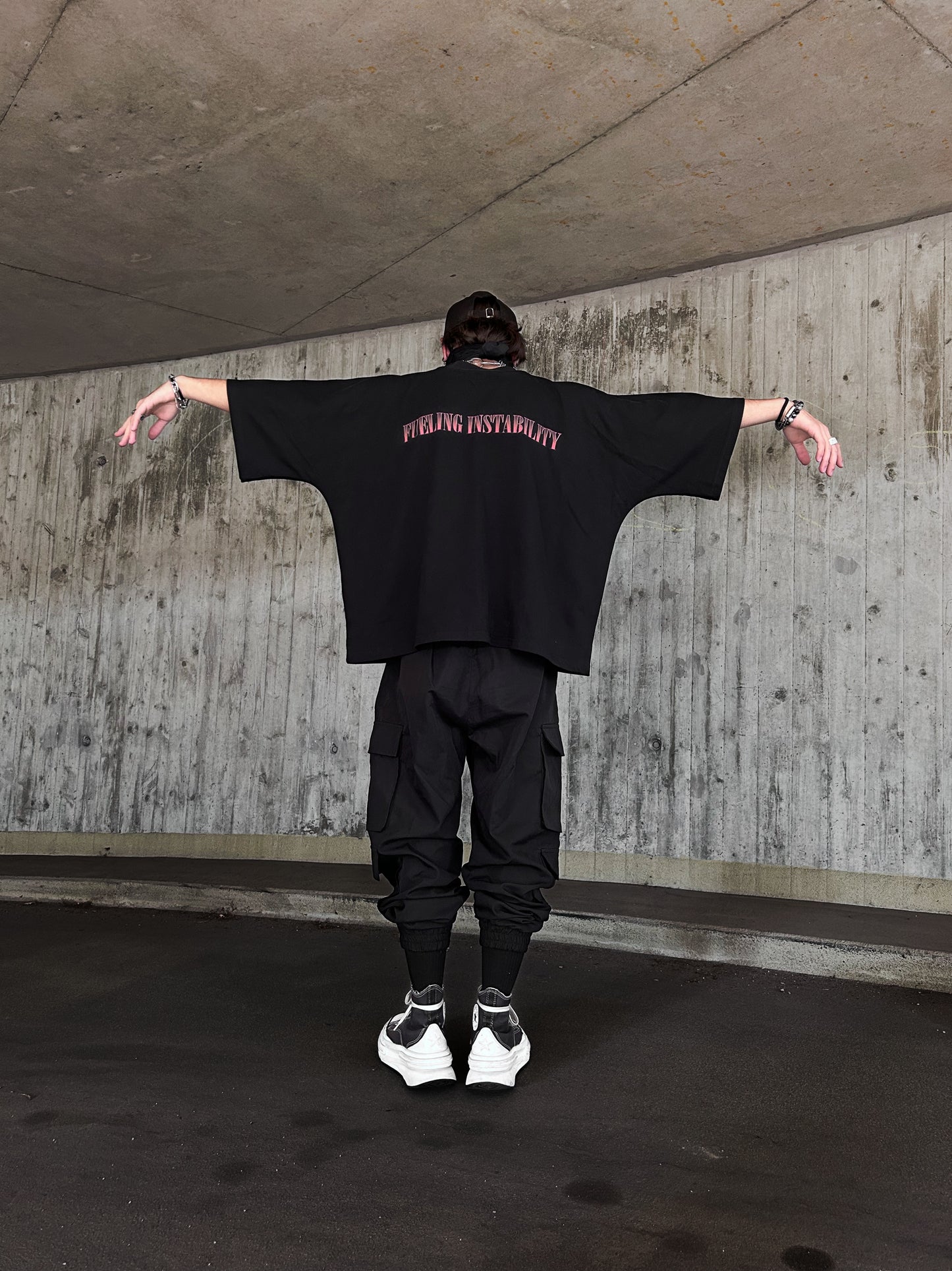 Person standing in front of a concrete wall wearing an oversized black t-shirt with ‘FUELING INSTABILITY’ text on the back, black cargo pants, and white sneakers. The person is facing away from the camera with arms outstretched, in an urban setting. The T-Shirt is by the brand SXDGXDS